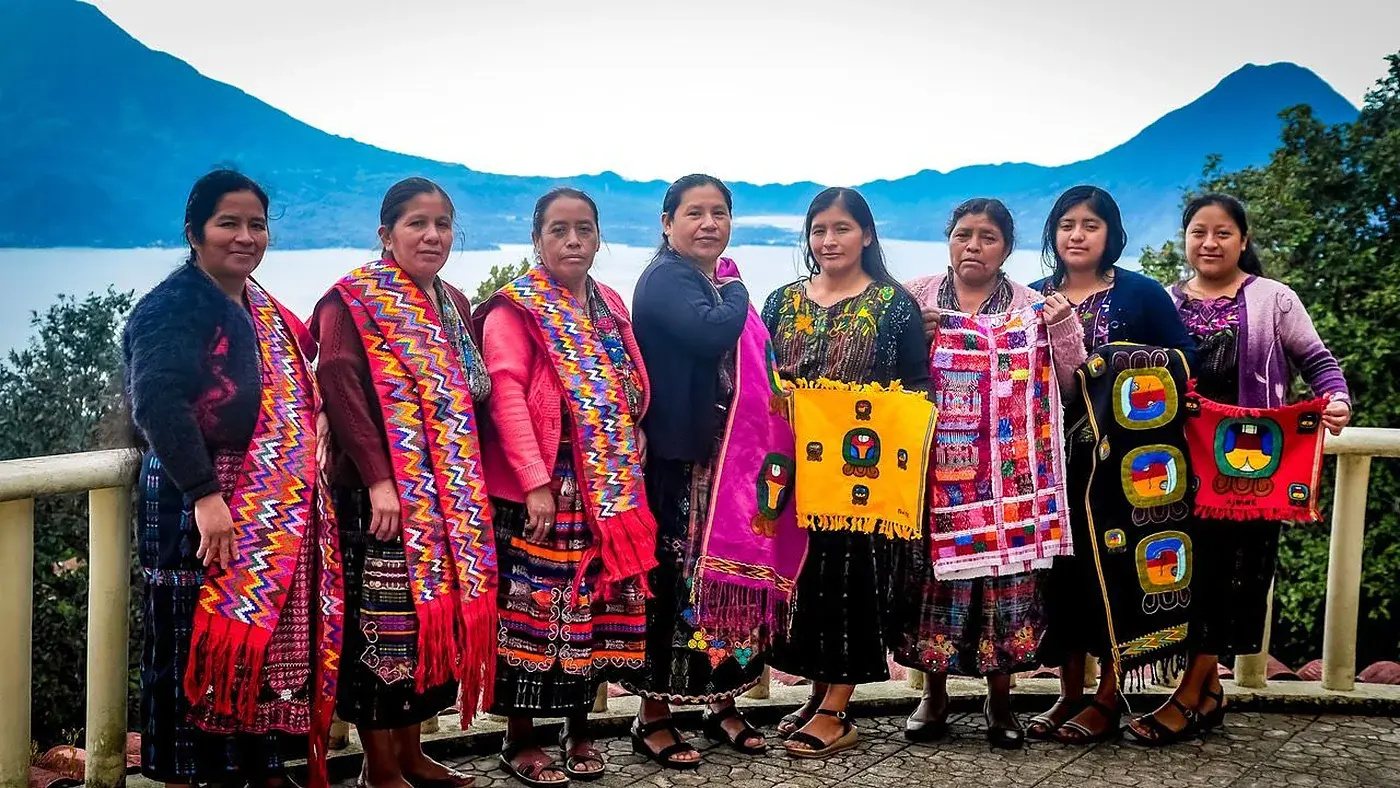 Eine Gruppe Frauen in traditioneller Kleidung zeigt ihre Webearbeiten, dahinter der See